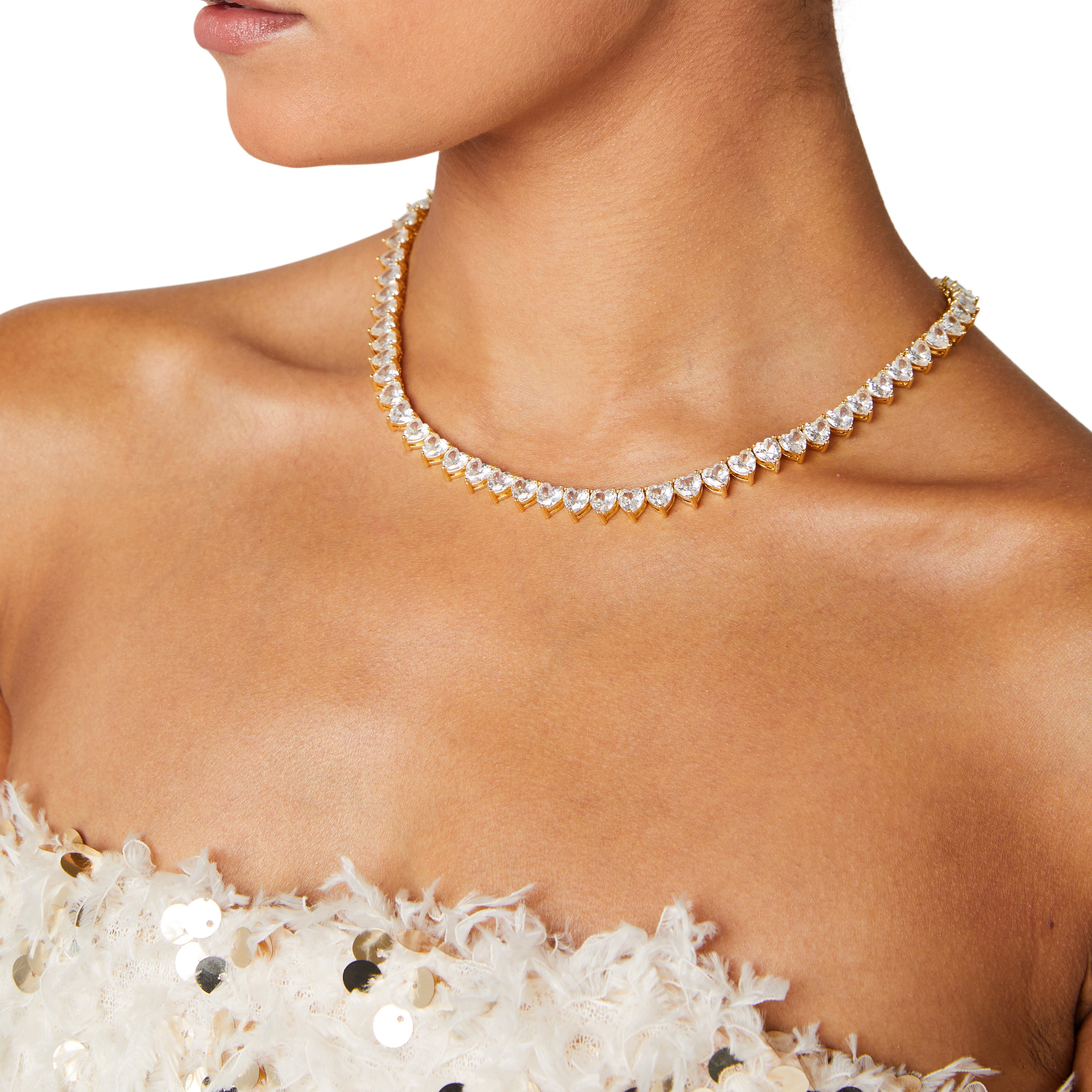 A deluxe tennis-style necklace composed of 7 mm heart-shaped clear cubic zirconia stones, each set in polished 18 ct gold plating. The hearts are aligned closely along a 16‑inch (40 cm) chain, creating a continuous sparkle around the neckline. The design features a secure, invisible clasp hidden behind one stone for a seamless look. The high-reflective gold finish and perfectly cut CZs give the piece a luxurious, radiant appeal—ideal for special occasions or elevating everyday style. It weighs approximately