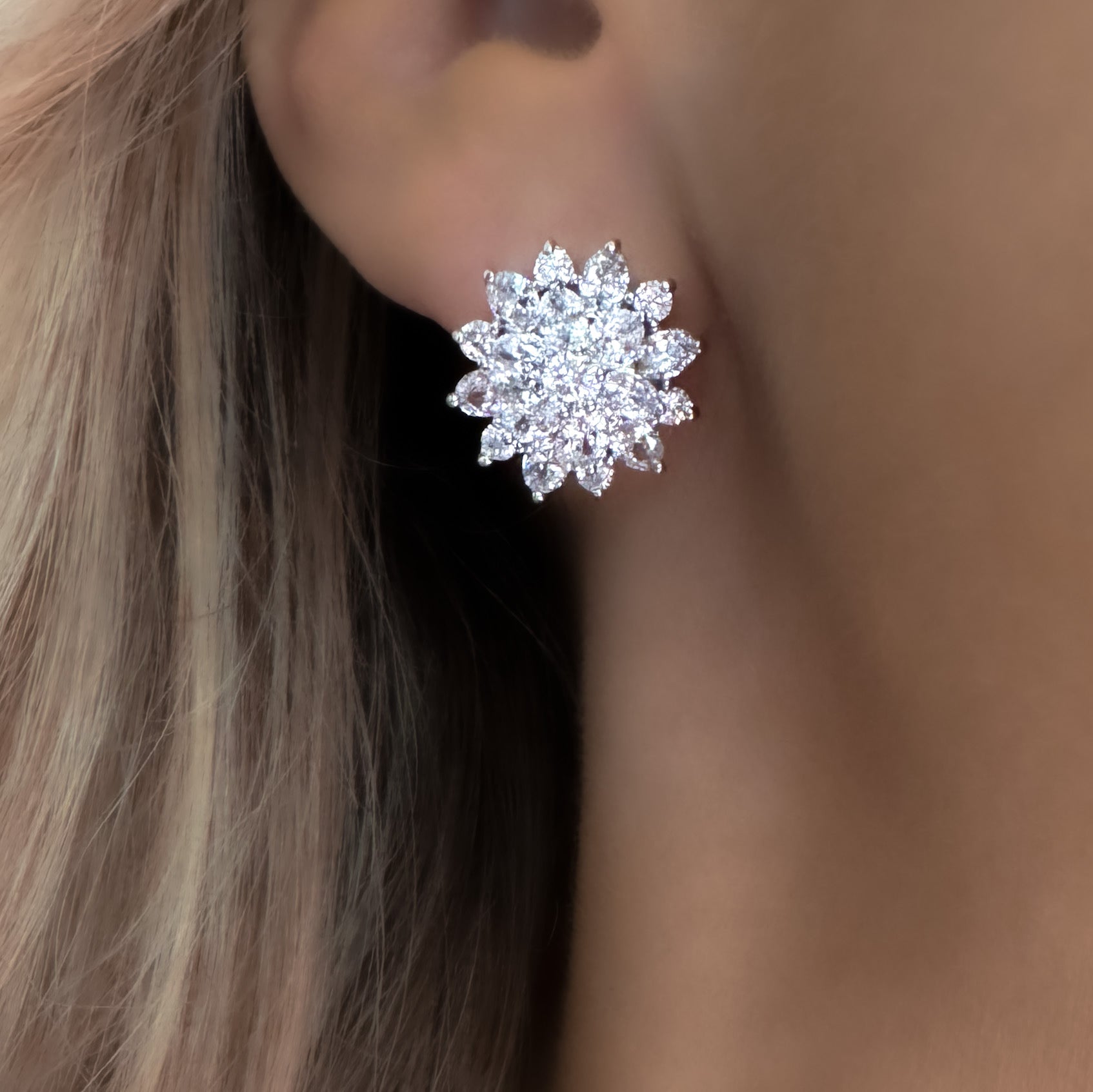 A pair of silver-plated stud earrings arranged side by side on a white background. Each earring is shaped like a stylised flower, featuring a central cluster of round-cut, brilliant cubic zirconia stones. Surrounding the center are two layers of sparkling CZ: an inner ring of smaller round stones, followed by an outer ring of marquise-shaped stones forming petals. The earrings measure approximately 1.6 cm in diameter and reflect light with a lustrous, glittering effect.