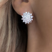 A pair of silver-plated stud earrings arranged side by side on a white background. Each earring is shaped like a stylised flower, featuring a central cluster of round-cut, brilliant cubic zirconia stones. Surrounding the center are two layers of sparkling CZ: an inner ring of smaller round stones, followed by an outer ring of marquise-shaped stones forming petals. The earrings measure approximately 1.6 cm in diameter and reflect light with a lustrous, glittering effect.