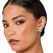 A pair of polished rhodium‑plated silver stud earrings shaped like stylised ribbon bows. Each bow features two gracefully looping ribbon forms with gently tapered tails that curve organically. The surface is smooth and reflective, catching light with soft gleams. The bows measure approximately 1.2 cm wide and 1 cm tall, and secure with standard butterfly push‑back posts. The design exudes charming femininity and understated elegance—a delicate accessory perfect for both everyday wear and subtle evening spa