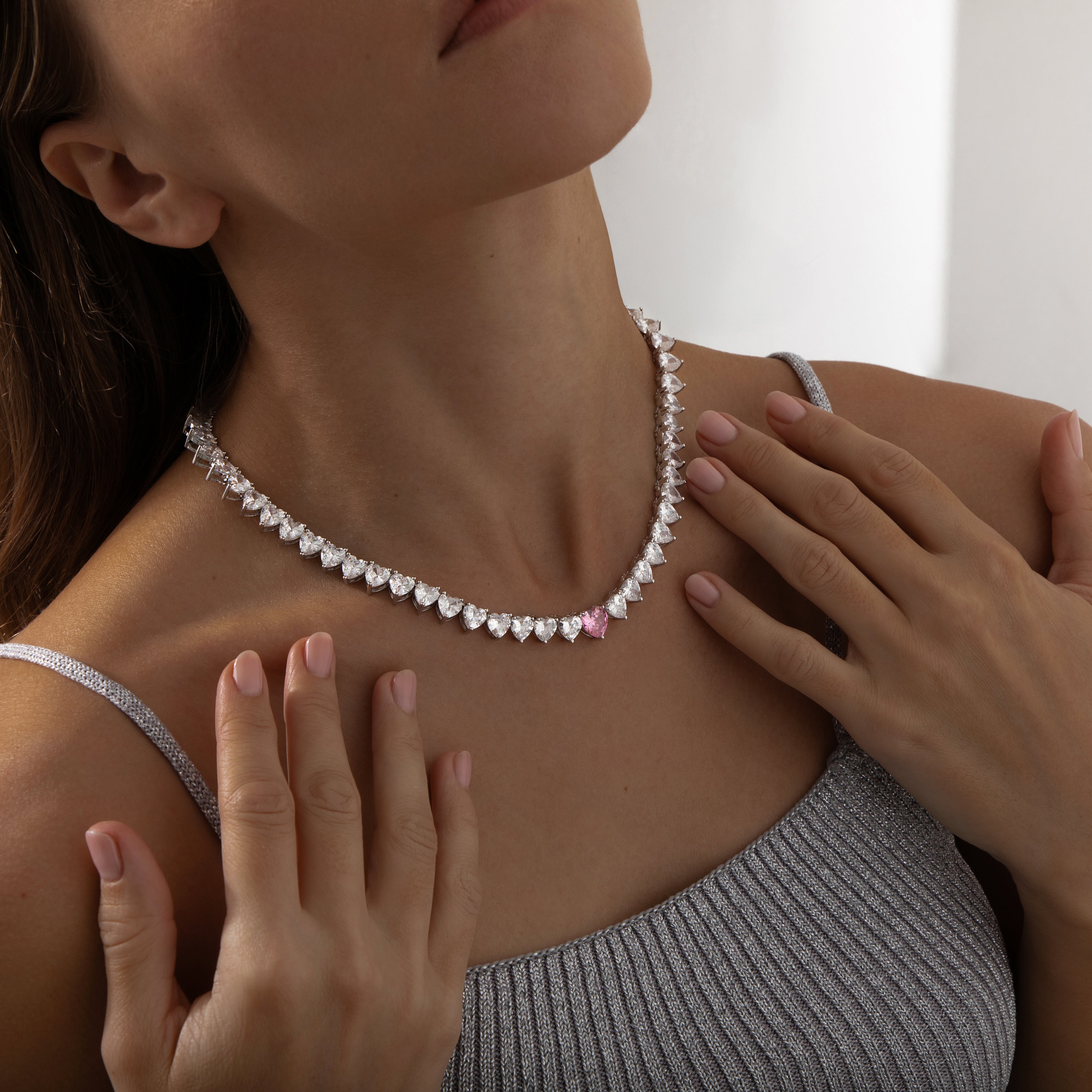 A sparkling silver-plated tennis-style necklace elegantly displayed on a soft, light background. It features a continuous row of heart-shaped cubic zirconia stones, each approximately 7 mm wide, set in rhodium-plated brass. The centerpiece is a single blush-pink heart stone (about 8 mm) that adds a pop of colour. The hearts are closely linked, creating a seamless chain around the neckline, and the full length is approximately 44 cm (17.5”) with a short extender. The polished silver setting enhances the bril