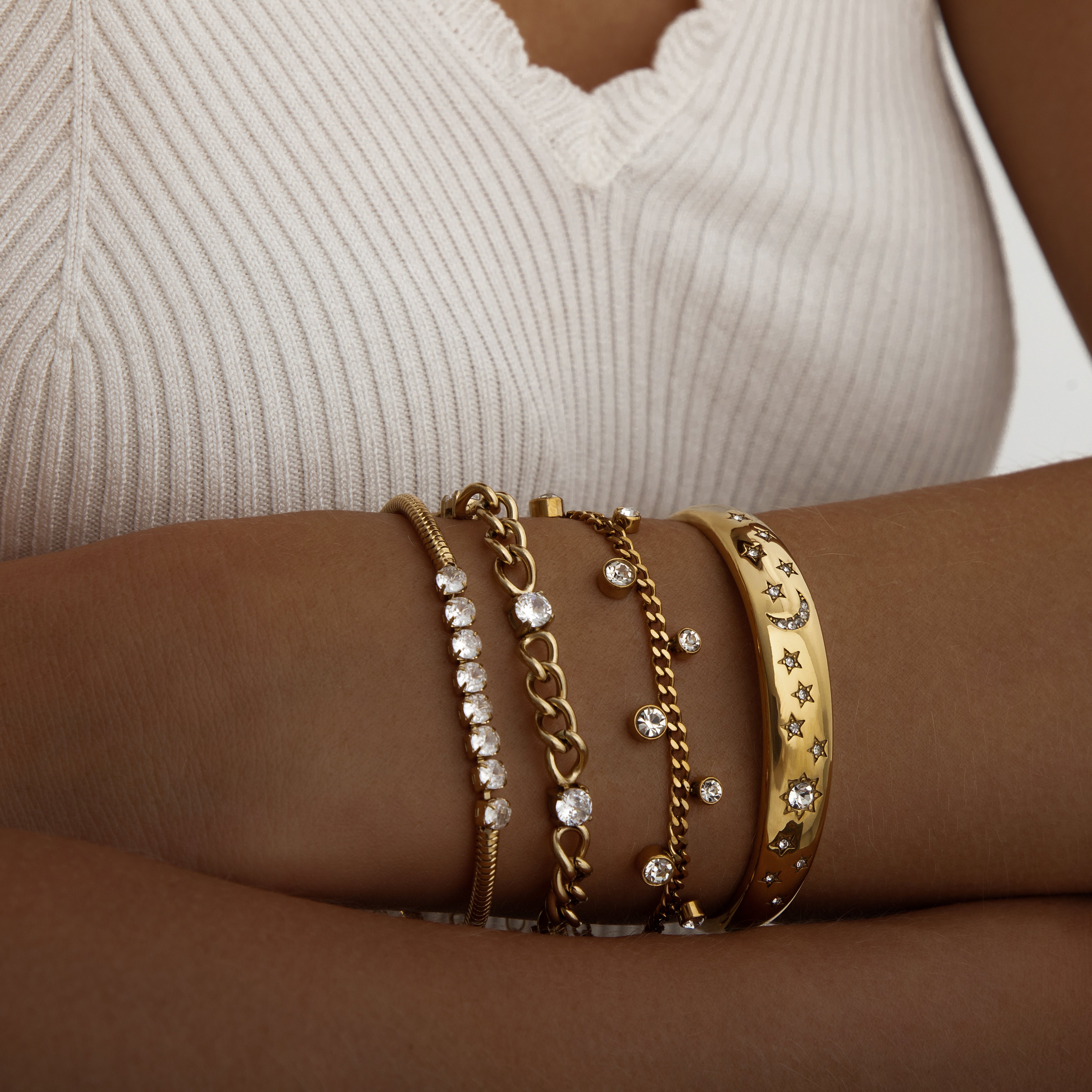 Gold stacking bracelets with star and chain designs on a wrist against a neutral background