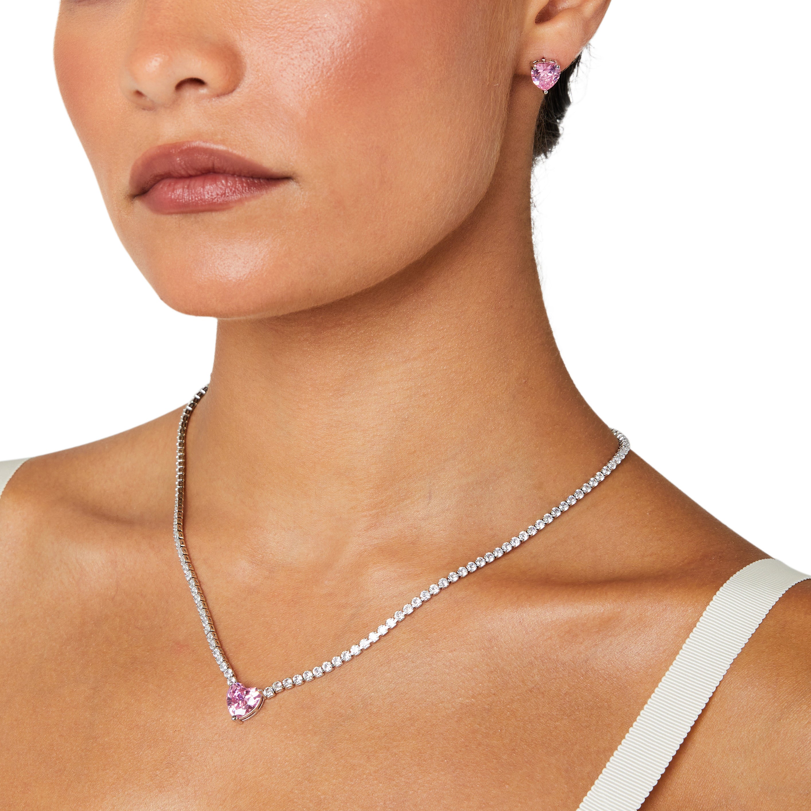 A matching set featuring a delicate silver-plated tennis‑style necklace and coordinating stud earrings displayed on a white background. The necklace consists of a continuous row of small, round white cubic zirconia stones (approximately 2–3 mm each), set in closely linked round bezels. At its centre hangs a single blush-pink, heart-shaped CZ pendant measuring about 8 mm across. Below the necklace, the set includes a pair of matching heart-shaped stud earrings in the same blush-pink tone, each about 8 mm dia