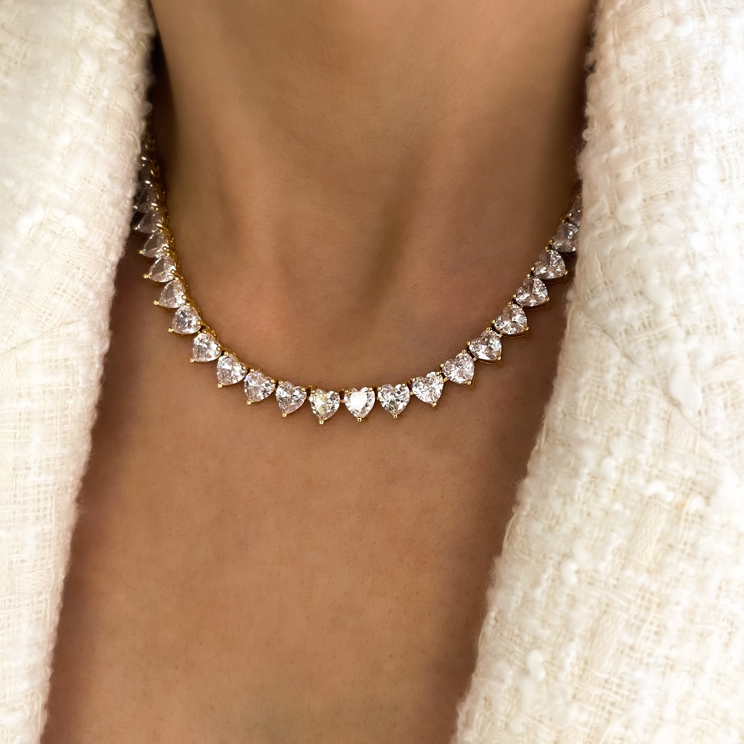 A deluxe tennis-style necklace composed of 7 mm heart-shaped clear cubic zirconia stones, each set in polished 18 ct gold plating. The hearts are aligned closely along a 16‑inch (40 cm) chain, creating a continuous sparkle around the neckline. The design features a secure, invisible clasp hidden behind one stone for a seamless look. The high-reflective gold finish and perfectly cut CZs give the piece a luxurious, radiant appeal—ideal for special occasions or elevating everyday style. It weighs approximately