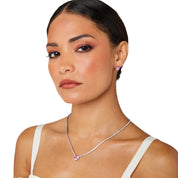 A matching set featuring a delicate silver-plated tennis‑style necklace and coordinating stud earrings displayed on a white background. The necklace consists of a continuous row of small, round white cubic zirconia stones (approximately 2–3 mm each), set in closely linked round bezels. At its centre hangs a single blush-pink, heart-shaped CZ pendant measuring about 8 mm across. Below the necklace, the set includes a pair of matching heart-shaped stud earrings in the same blush-pink tone, each about 8 mm dia