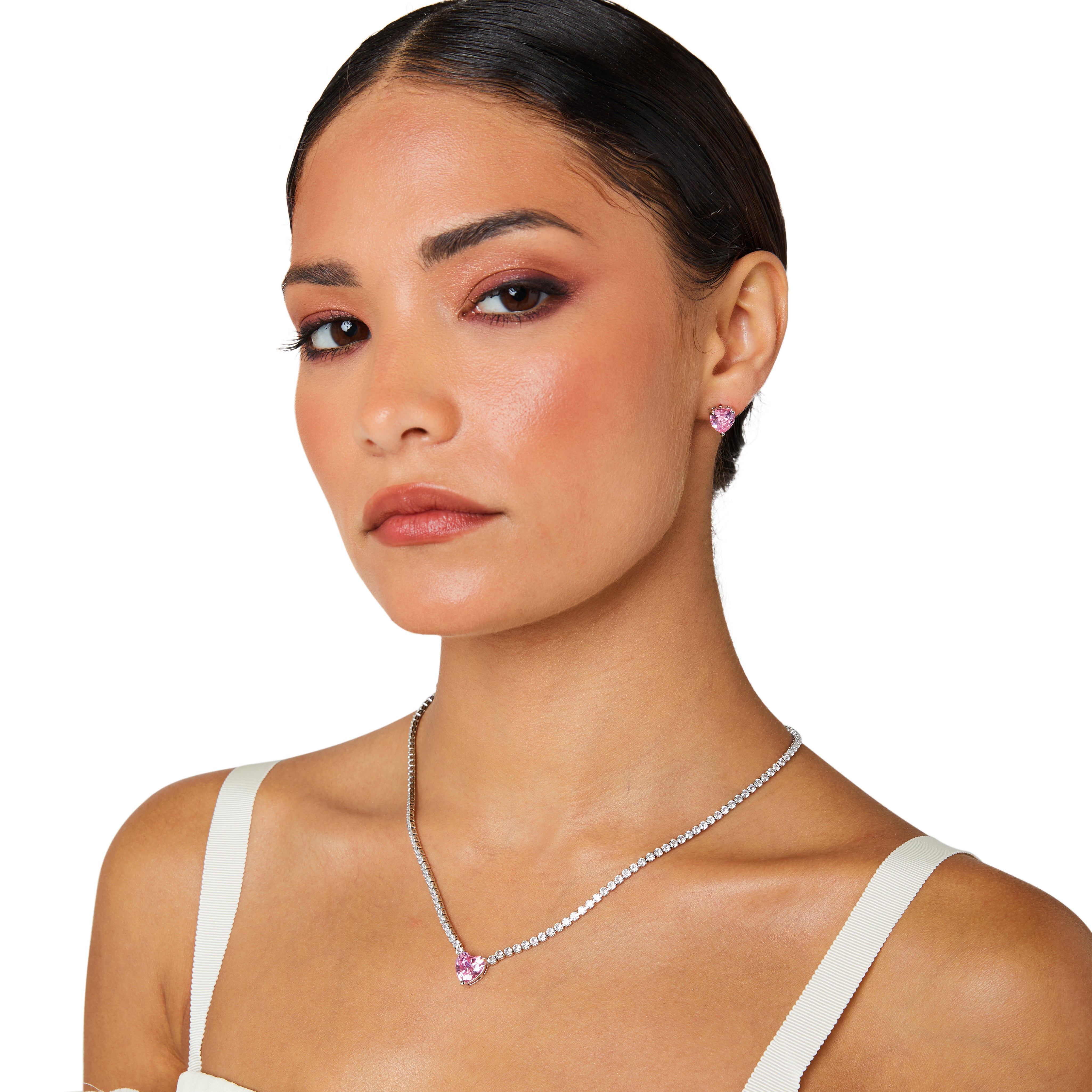 A matching set featuring a delicate silver-plated tennis‑style necklace and coordinating stud earrings displayed on a white background. The necklace consists of a continuous row of small, round white cubic zirconia stones (approximately 2–3 mm each), set in closely linked round bezels. At its centre hangs a single blush-pink, heart-shaped CZ pendant measuring about 8 mm across. Below the necklace, the set includes a pair of matching heart-shaped stud earrings in the same blush-pink tone, each about 8 mm dia