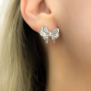 A pair of polished rhodium‑plated silver stud earrings shaped like stylised ribbon bows. Each bow features two gracefully looping ribbon forms with gently tapered tails that curve organically. The surface is smooth and reflective, catching light with soft gleams. The bows measure approximately 1.2 cm wide and 1 cm tall, and secure with standard butterfly push‑back posts. The design exudes charming femininity and understated elegance—a delicate accessory perfect for both everyday wear and subtle evening spa