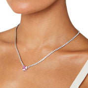 A matching set featuring a delicate silver-plated tennis‑style necklace and coordinating stud earrings displayed on a white background. The necklace consists of a continuous row of small, round white cubic zirconia stones (approximately 2–3 mm each), set in closely linked round bezels. At its centre hangs a single blush-pink, heart-shaped CZ pendant measuring about 8 mm across. Below the necklace, the set includes a pair of matching heart-shaped stud earrings in the same blush-pink tone, each about 8 mm dia