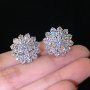 A pair of silver-plated stud earrings arranged side by side on a white background. Each earring is shaped like a stylised flower, featuring a central cluster of round-cut, brilliant cubic zirconia stones. Surrounding the center are two layers of sparkling CZ: an inner ring of smaller round stones, followed by an outer ring of marquise-shaped stones forming petals. The earrings measure approximately 1.6 cm in diameter and reflect light with a lustrous, glittering effect.