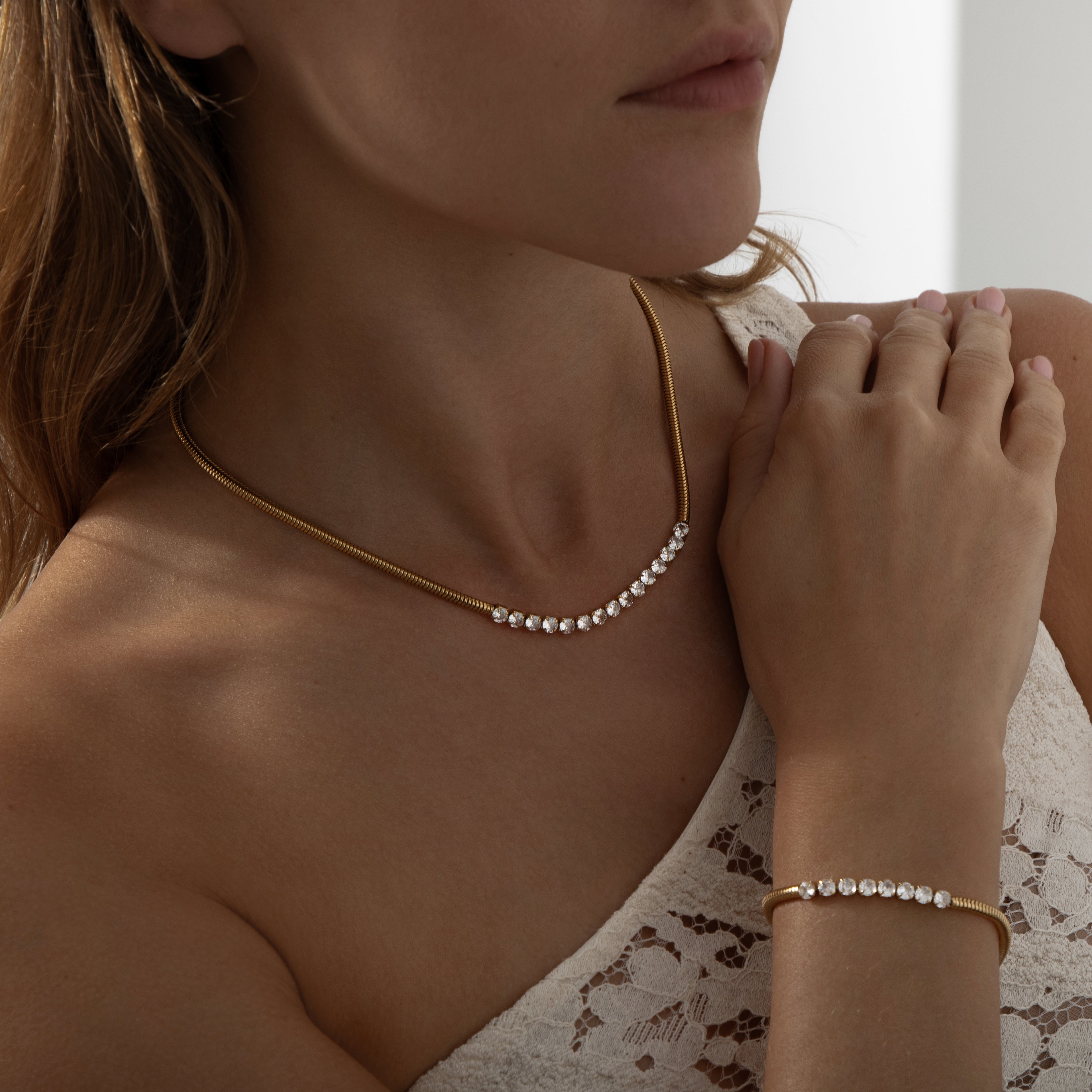 A layered-style necklace combining two distinct chains: one side is a classic slender snake chain, the other a sparkling row of cubic zirconia stones set in bezel-like settings (tennis chain). Both strands merge at a central point and rest together on the collarbone. The chains are 16K gold‑plated stainless steel, with the CZ strand approximately 4 mm wide. The necklace measures around 41‑46 cm, secured by a small lobster‑claw clasp with an extender. The warm gold finish contrasts the smooth metallic sheen