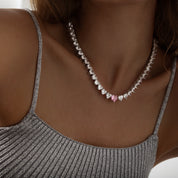 A sparkling silver-plated tennis-style necklace elegantly displayed on a soft, light background. It features a continuous row of heart-shaped cubic zirconia stones, each approximately 7 mm wide, set in rhodium-plated brass. The centerpiece is a single blush-pink heart stone (about 8 mm) that adds a pop of colour. The hearts are closely linked, creating a seamless chain around the neckline, and the full length is approximately 44 cm (17.5”) with a short extender. The polished silver setting enhances the bril