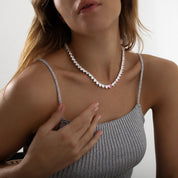 A sparkling silver-plated tennis-style necklace elegantly displayed on a soft, light background. It features a continuous row of heart-shaped cubic zirconia stones, each approximately 7 mm wide, set in rhodium-plated brass. The centerpiece is a single blush-pink heart stone (about 8 mm) that adds a pop of colour. The hearts are closely linked, creating a seamless chain around the neckline, and the full length is approximately 44 cm (17.5”) with a short extender. The polished silver setting enhances the bril