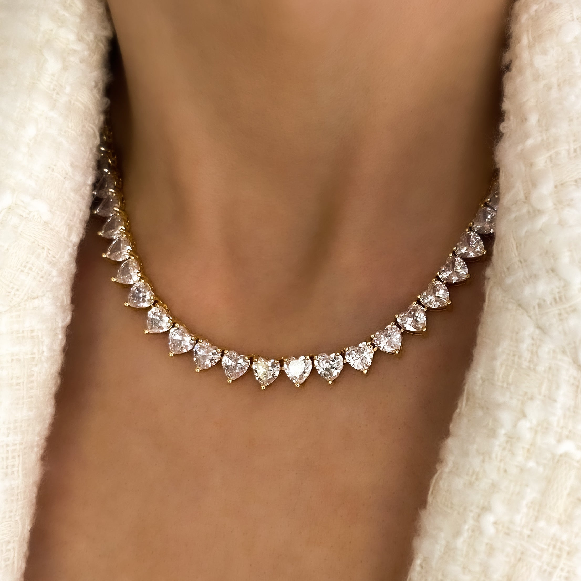 A deluxe tennis-style necklace composed of 7 mm heart-shaped clear cubic zirconia stones, each set in polished 18 ct gold plating. The hearts are aligned closely along a 16‑inch (40 cm) chain, creating a continuous sparkle around the neckline. The design features a secure, invisible clasp hidden behind one stone for a seamless look. The high-reflective gold finish and perfectly cut CZs give the piece a luxurious, radiant appeal—ideal for special occasions or elevating everyday style. It weighs approximately
