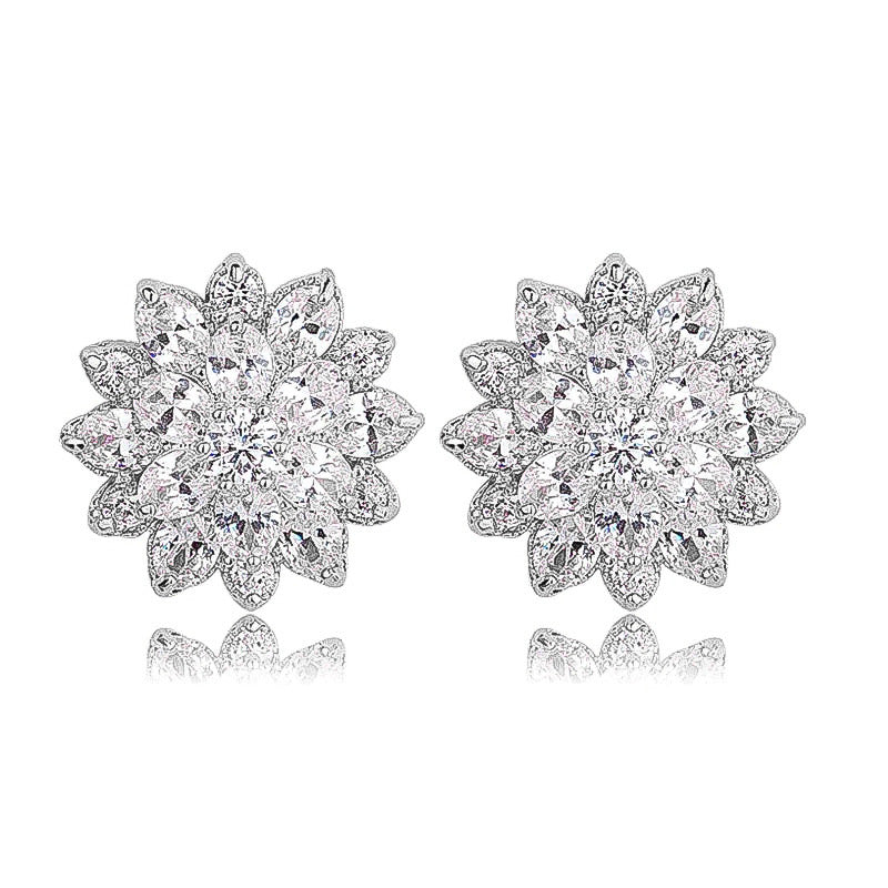 A pair of silver-plated stud earrings arranged side by side on a white background. Each earring is shaped like a stylised flower, featuring a central cluster of round-cut, brilliant cubic zirconia stones. Surrounding the center are two layers of sparkling CZ: an inner ring of smaller round stones, followed by an outer ring of marquise-shaped stones forming petals. The earrings measure approximately 1.6 cm in diameter and reflect light with a lustrous, glittering effect.