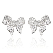 A pair of polished rhodium‑plated silver stud earrings shaped like stylised ribbon bows. Each bow features two gracefully looping ribbon forms with gently tapered tails that curve organically. The surface is smooth and reflective, catching light with soft gleams. The bows measure approximately 1.2 cm wide and 1 cm tall, and secure with standard butterfly push‑back posts. The design exudes charming femininity and understated elegance—a delicate accessory perfect for both everyday wear and subtle evening spa