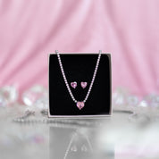 A matching set featuring a delicate silver-plated tennis‑style necklace and coordinating stud earrings displayed on a white background. The necklace consists of a continuous row of small, round white cubic zirconia stones (approximately 2–3 mm each), set in closely linked round bezels. At its centre hangs a single blush-pink, heart-shaped CZ pendant measuring about 8 mm across. Below the necklace, the set includes a pair of matching heart-shaped stud earrings in the same blush-pink tone, each about 8 mm dia