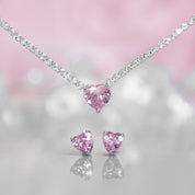 A matching set featuring a delicate silver-plated tennis‑style necklace and coordinating stud earrings displayed on a white background. The necklace consists of a continuous row of small, round white cubic zirconia stones (approximately 2–3 mm each), set in closely linked round bezels. At its centre hangs a single blush-pink, heart-shaped CZ pendant measuring about 8 mm across. Below the necklace, the set includes a pair of matching heart-shaped stud earrings in the same blush-pink tone, each about 8 mm dia