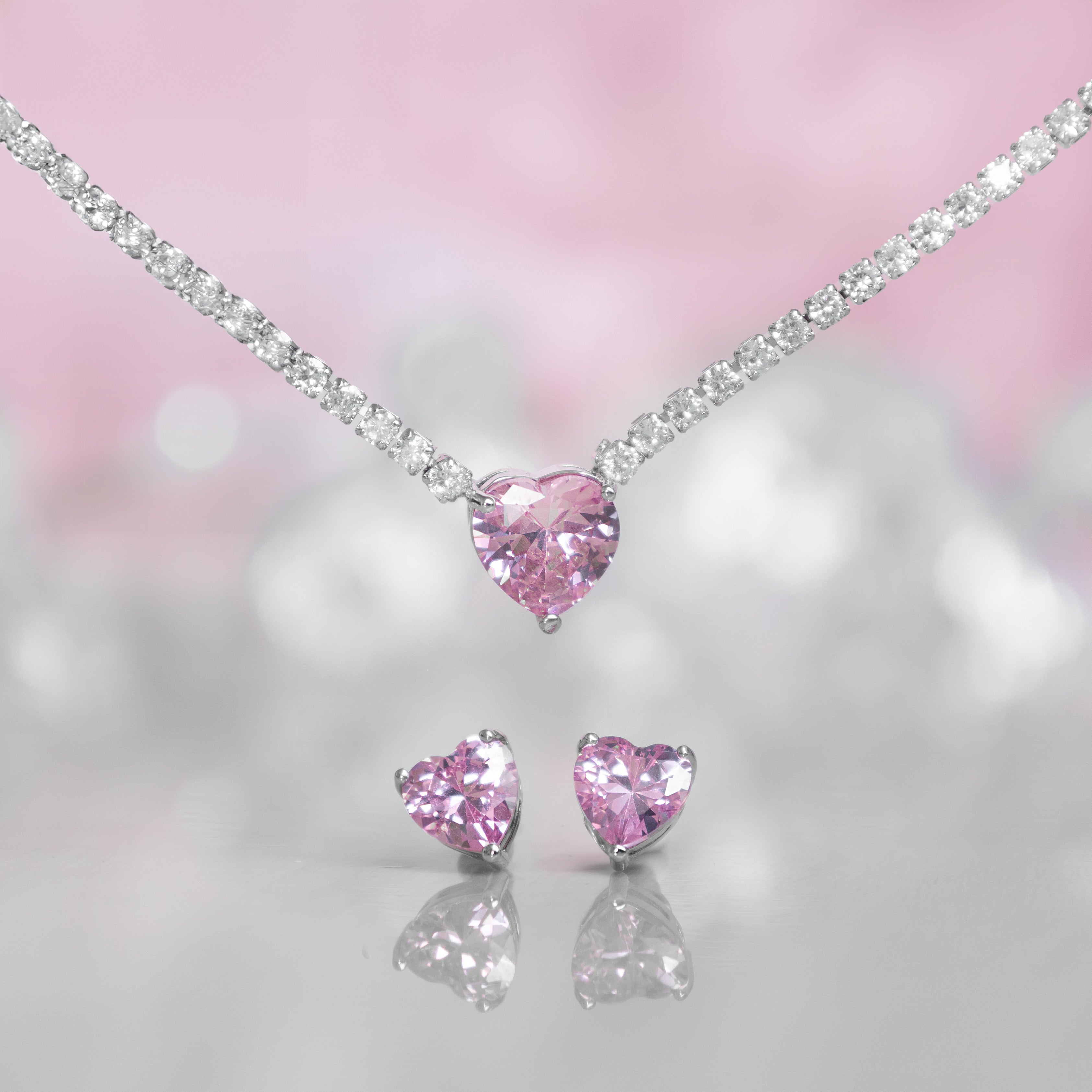 A matching set featuring a delicate silver-plated tennis‑style necklace and coordinating stud earrings displayed on a white background. The necklace consists of a continuous row of small, round white cubic zirconia stones (approximately 2–3 mm each), set in closely linked round bezels. At its centre hangs a single blush-pink, heart-shaped CZ pendant measuring about 8 mm across. Below the necklace, the set includes a pair of matching heart-shaped stud earrings in the same blush-pink tone, each about 8 mm dia