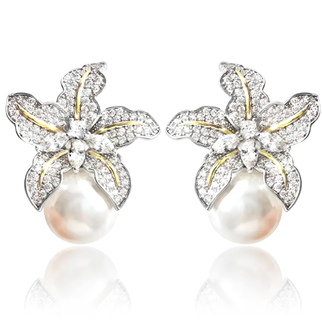 A pair of dangling earrings photographed against a neutral, softly lit background. Each earring features a tiny wild iris flower crafted in rhodium-plated silver, with sculpted petals that fan outward and slightly curl at the edges. Below the flower hangs a luminous baroque freshwater pearl—irregular in shape—with a creamy, iridescent surface that catches subtle tones of pink and white. The earrings measure approximately 2.3 cm from top of the flower to the base of the pearl and fasten with hypoallergenic p