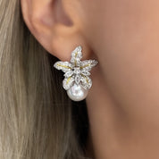 A pair of dangling earrings photographed against a neutral, softly lit background. Each earring features a tiny wild iris flower crafted in rhodium-plated silver, with sculpted petals that fan outward and slightly curl at the edges. Below the flower hangs a luminous baroque freshwater pearl—irregular in shape—with a creamy, iridescent surface that catches subtle tones of pink and white. The earrings measure approximately 2.3 cm from top of the flower to the base of the pearl and fasten with hypoallergenic p