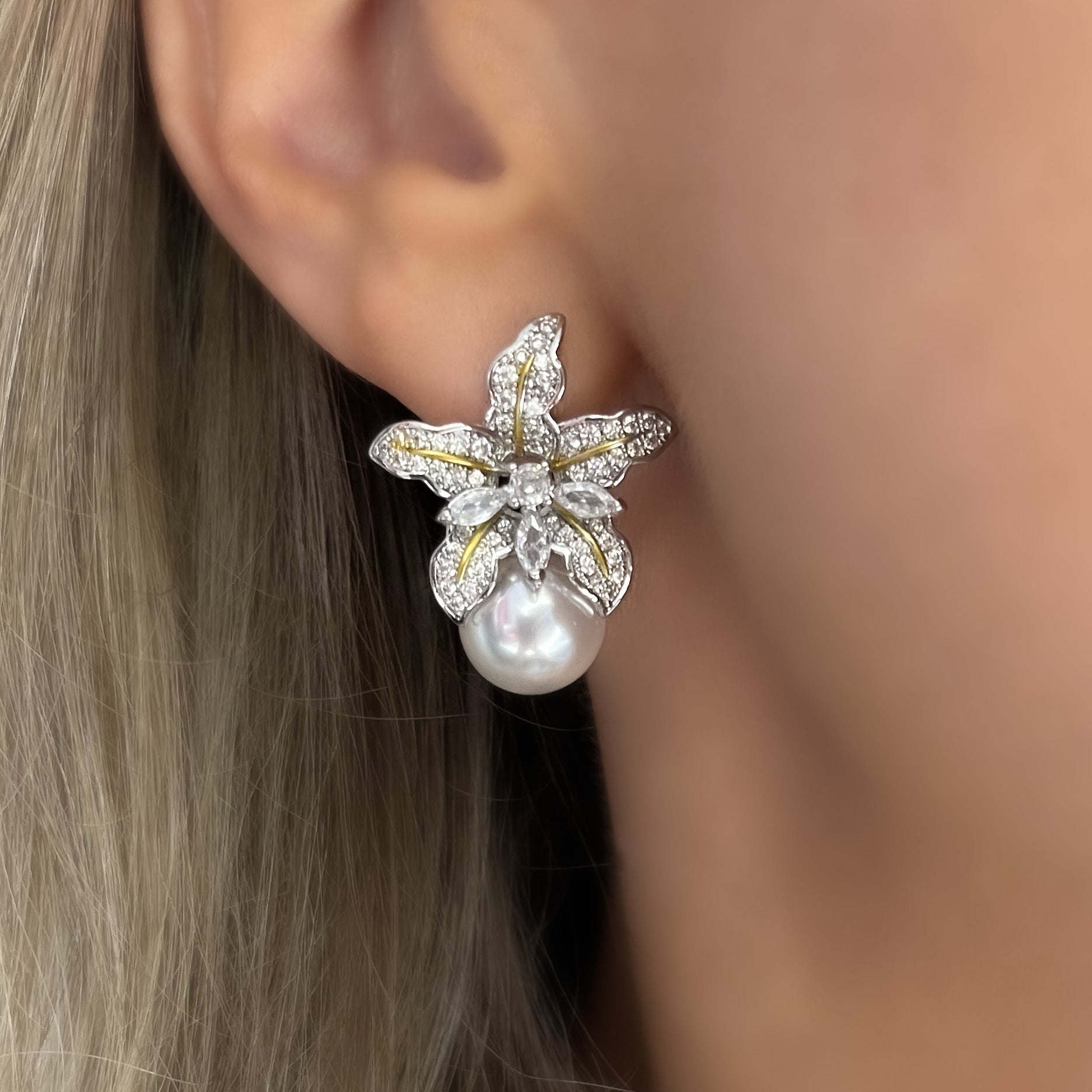 A pair of dangling earrings photographed against a neutral, softly lit background. Each earring features a tiny wild iris flower crafted in rhodium-plated silver, with sculpted petals that fan outward and slightly curl at the edges. Below the flower hangs a luminous baroque freshwater pearl—irregular in shape—with a creamy, iridescent surface that catches subtle tones of pink and white. The earrings measure approximately 2.3 cm from top of the flower to the base of the pearl and fasten with hypoallergenic p
