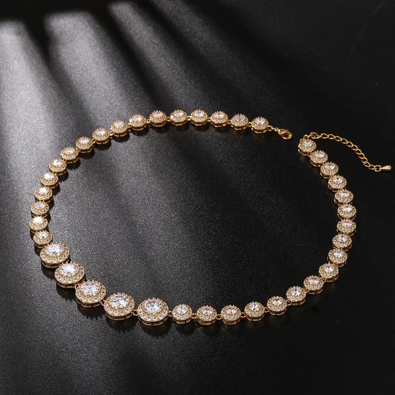 gold crystal tennis necklace with graduated cubic zirconia stones and pavé halos, photographed against a black background to highlight the necklace’s radiant sparkle and elegant design