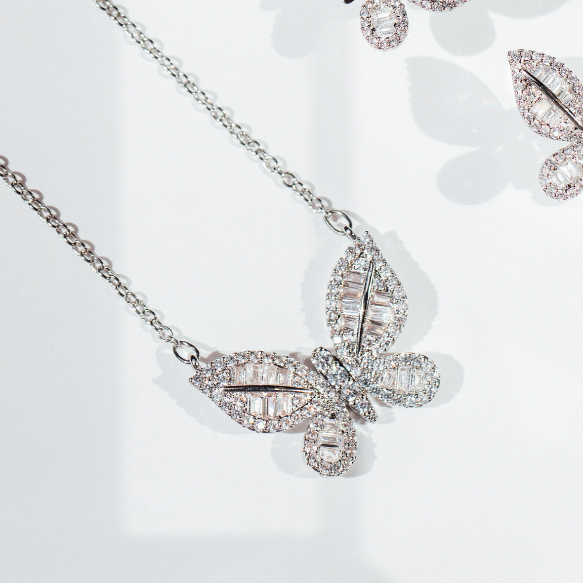 A delicately crafted rhodium-plated silver cable chain suspends a butterfly pendant measuring about 1.8 cm across. The butterfly’s wings are outlined and filled with sparkling white cubic zirconia crystals of varying cuts—round and baguette—creating a textured, shimmering mosaic. The pendant is centred on the chain, which measures 16 in (40 cm) and fastens with a small spring-ring clasp. The overall aesthetic is elegant, glimmering, and nature-inspired, perfect for adding a subtle yet eye-catching accent to