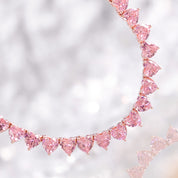 A rose‑gold plated tennis‑style necklace drapes elegantly around a model’s neck. The piece features a continuous row of blush‑pink, heart‑shaped cubic zirconia stones, each approximately 5 mm wide, linked closely together. The necklace measures about 17 in (43 cm) with a 0.5 in extender and fastens with a secure clasp. The polished rose‑gold setting enhances the sparkle of the pink hearts, creating a romantic, feminine shimmer perfect for both everyday wear and special occasions.