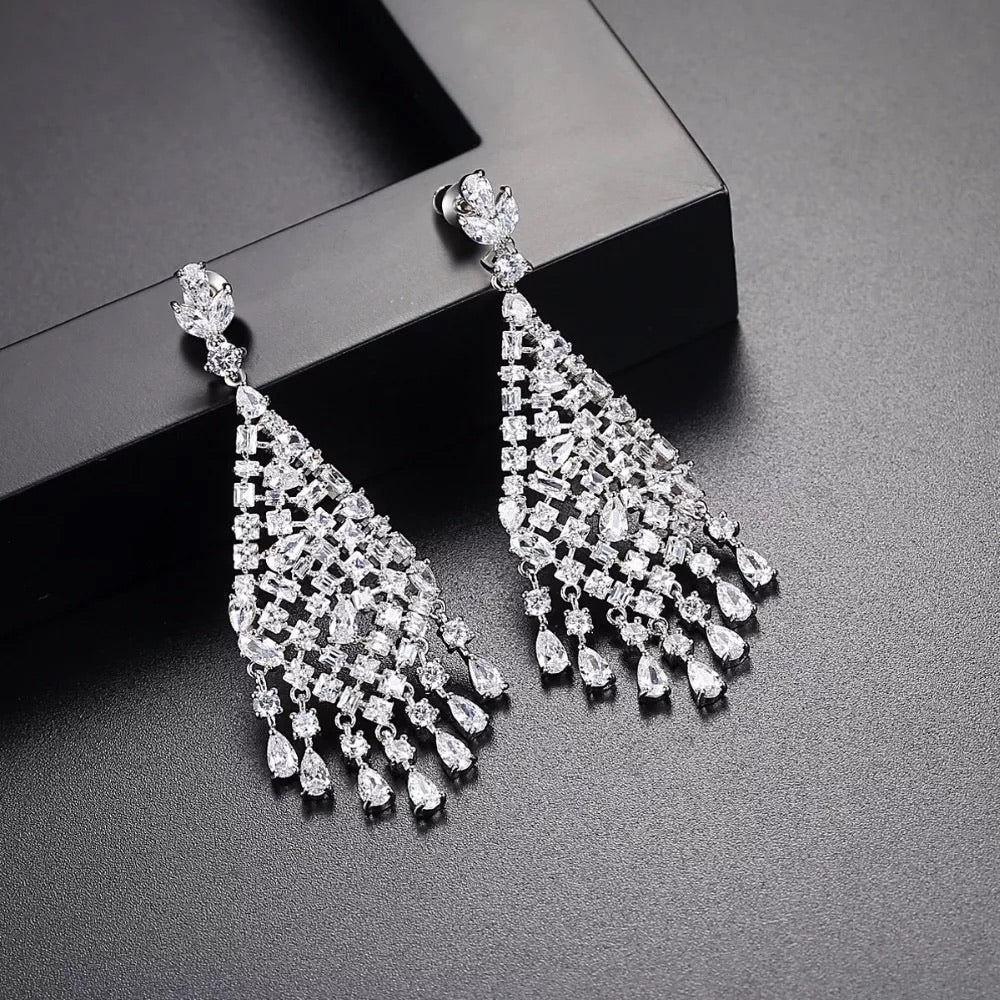 Silver chandelier earrings featuring cascading white cubic zirconia stones, rhodium-plated with a 6.4 cm drop, shown on model and against light and dark backgrounds.