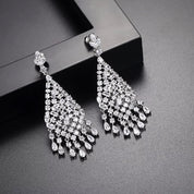 Silver chandelier earrings featuring cascading white cubic zirconia stones, rhodium-plated with a 6.4 cm drop, shown on model and against light and dark backgrounds.