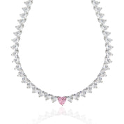 A sparkling silver-plated tennis-style necklace elegantly displayed on a soft, light background. It features a continuous row of heart-shaped cubic zirconia stones, each approximately 7 mm wide, set in rhodium-plated brass. The centerpiece is a single blush-pink heart stone (about 8 mm) that adds a pop of colour. The hearts are closely linked, creating a seamless chain around the neckline, and the full length is approximately 44 cm (17.5”) with a short extender. The polished silver setting enhances the bril