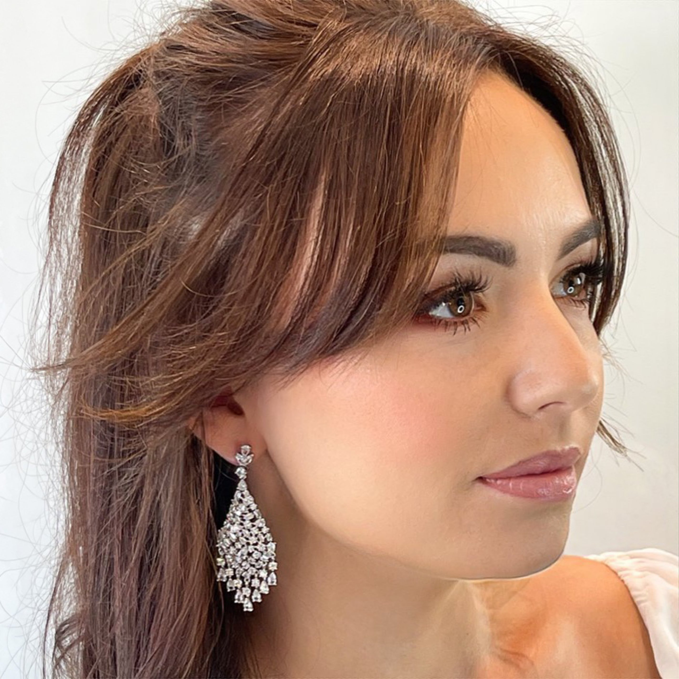 Silver chandelier earrings featuring cascading white cubic zirconia stones, rhodium-plated with a 6.4 cm drop, shown on model and against light and dark backgrounds.