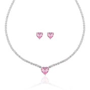 A matching set featuring a delicate silver-plated tennis‑style necklace and coordinating stud earrings displayed on a white background. The necklace consists of a continuous row of small, round white cubic zirconia stones (approximately 2–3 mm each), set in closely linked round bezels. At its centre hangs a single blush-pink, heart-shaped CZ pendant measuring about 8 mm across. Below the necklace, the set includes a pair of matching heart-shaped stud earrings in the same blush-pink tone, each about 8 mm dia