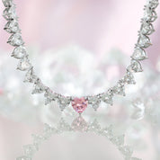 A sparkling silver-plated tennis-style necklace elegantly displayed on a soft, light background. It features a continuous row of heart-shaped cubic zirconia stones, each approximately 7 mm wide, set in rhodium-plated brass. The centerpiece is a single blush-pink heart stone (about 8 mm) that adds a pop of colour. The hearts are closely linked, creating a seamless chain around the neckline, and the full length is approximately 44 cm (17.5”) with a short extender. The polished silver setting enhances the bril