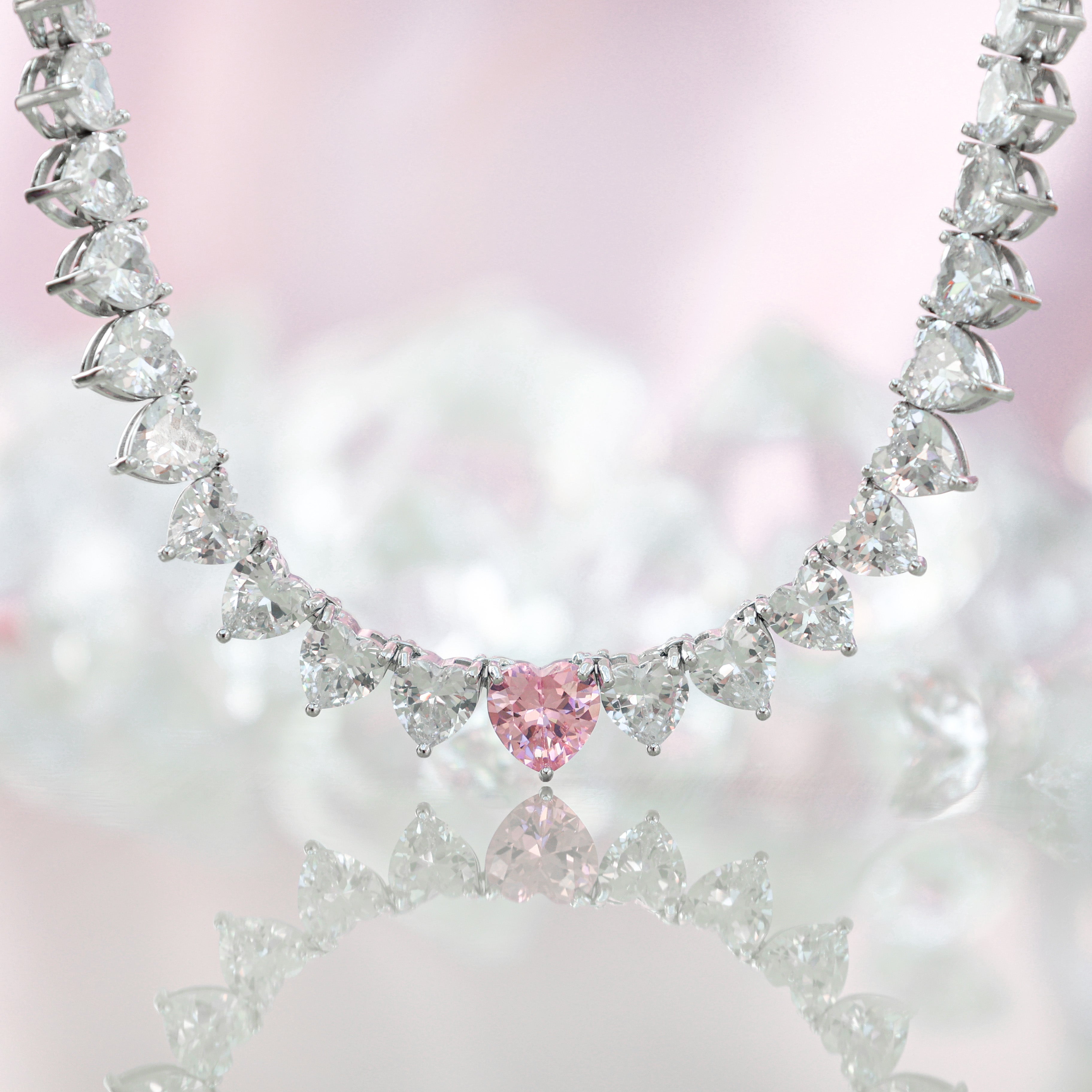 A sparkling silver-plated tennis-style necklace elegantly displayed on a soft, light background. It features a continuous row of heart-shaped cubic zirconia stones, each approximately 7 mm wide, set in rhodium-plated brass. The centerpiece is a single blush-pink heart stone (about 8 mm) that adds a pop of colour. The hearts are closely linked, creating a seamless chain around the neckline, and the full length is approximately 44 cm (17.5”) with a short extender. The polished silver setting enhances the bril
