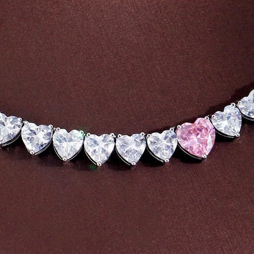 A sparkling silver-plated tennis-style necklace elegantly displayed on a soft, light background. It features a continuous row of heart-shaped cubic zirconia stones, each approximately 7 mm wide, set in rhodium-plated brass. The centerpiece is a single blush-pink heart stone (about 8 mm) that adds a pop of colour. The hearts are closely linked, creating a seamless chain around the neckline, and the full length is approximately 44 cm (17.5”) with a short extender. The polished silver setting enhances the bril