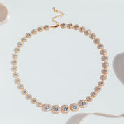 gold crystal tennis necklace with graduated cubic zirconia stones and pavé halos, photographed against a white background to highlight the necklace’s radiant sparkle and elegant design