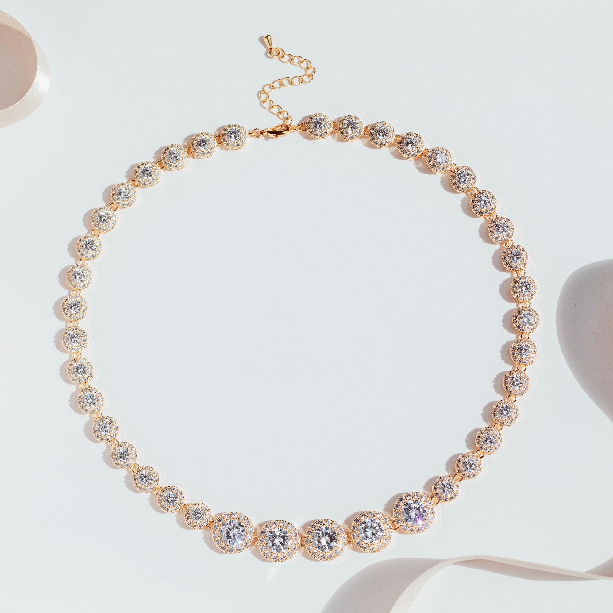 gold crystal tennis necklace with graduated cubic zirconia stones and pavé halos, photographed against a white background to highlight the necklace’s radiant sparkle and elegant design