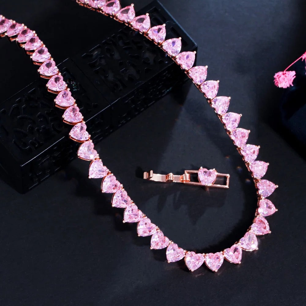 A rose‑gold plated tennis‑style necklace drapes elegantly around a model’s neck. The piece features a continuous row of blush‑pink, heart‑shaped cubic zirconia stones, each approximately 5 mm wide, linked closely together. The necklace measures about 17 in (43 cm) with a 0.5 in extender and fastens with a secure clasp. The polished rose‑gold setting enhances the sparkle of the pink hearts, creating a romantic, feminine shimmer perfect for both everyday wear and special occasions.