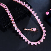 A rose‑gold plated tennis‑style necklace drapes elegantly around a model’s neck. The piece features a continuous row of blush‑pink, heart‑shaped cubic zirconia stones, each approximately 5 mm wide, linked closely together. The necklace measures about 17 in (43 cm) with a 0.5 in extender and fastens with a secure clasp. The polished rose‑gold setting enhances the sparkle of the pink hearts, creating a romantic, feminine shimmer perfect for both everyday wear and special occasions.