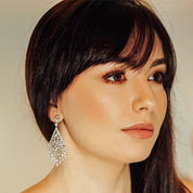 Silver chandelier earrings featuring cascading white cubic zirconia stones, rhodium-plated with a 6.4 cm drop, shown on model and against light and dark backgrounds.