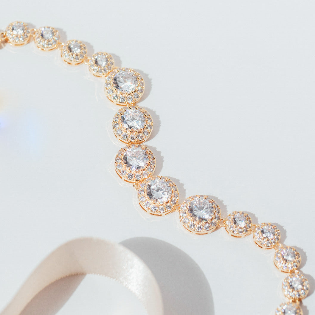 gold crystal tennis necklace with graduated cubic zirconia stones and pavé halos, photographed against a white background to highlight the necklace’s radiant sparkle and elegant design