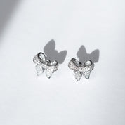 A pair of polished rhodium‑plated silver stud earrings shaped like stylised ribbon bows. Each bow features two gracefully looping ribbon forms with gently tapered tails that curve organically. The surface is smooth and reflective, catching light with soft gleams. The bows measure approximately 1.2 cm wide and 1 cm tall, and secure with standard butterfly push‑back posts. The design exudes charming femininity and understated elegance—a delicate accessory perfect for both everyday wear and subtle evening spa
