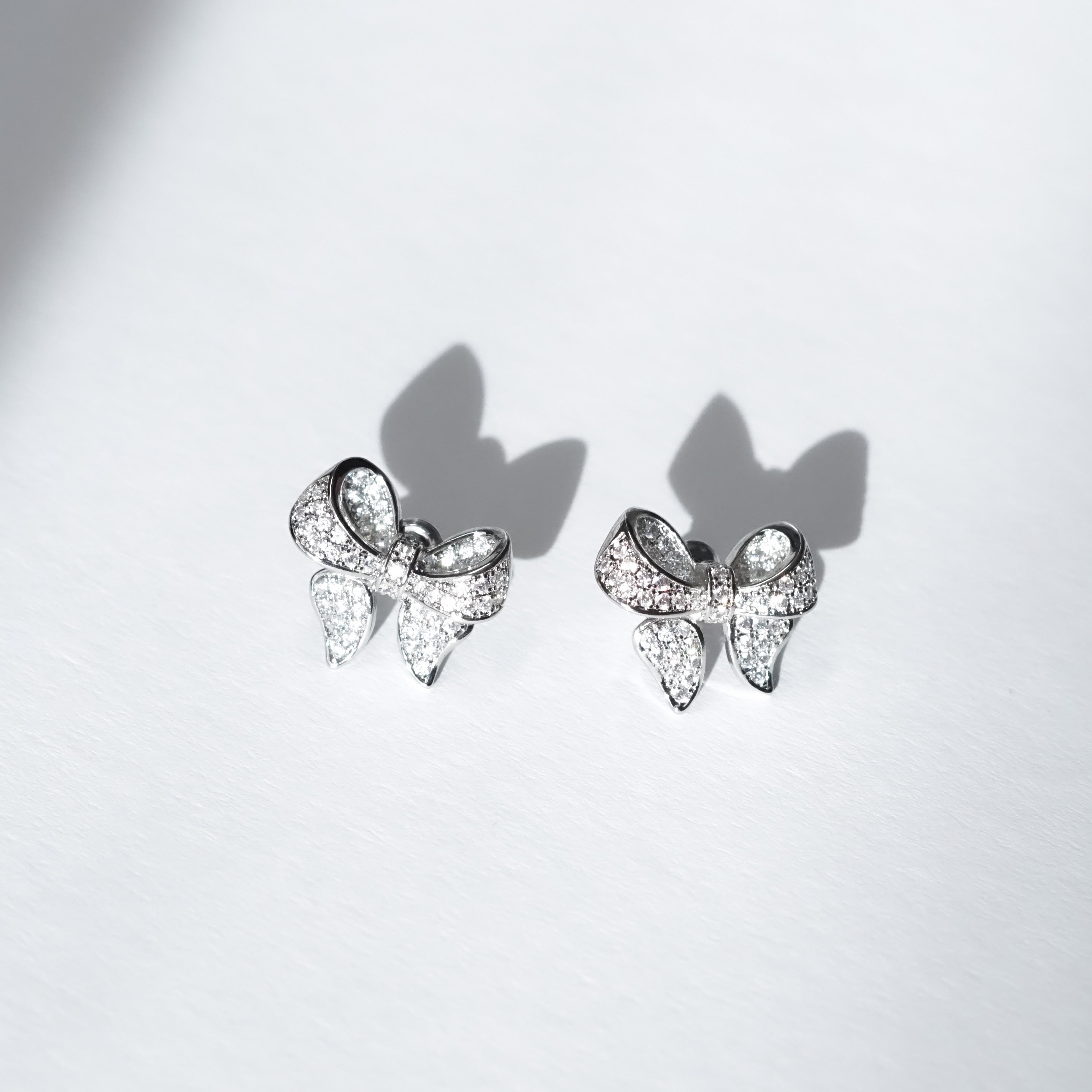 A pair of polished rhodium‑plated silver stud earrings shaped like stylised ribbon bows. Each bow features two gracefully looping ribbon forms with gently tapered tails that curve organically. The surface is smooth and reflective, catching light with soft gleams. The bows measure approximately 1.2 cm wide and 1 cm tall, and secure with standard butterfly push‑back posts. The design exudes charming femininity and understated elegance—a delicate accessory perfect for both everyday wear and subtle evening spa