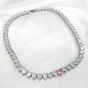 A sparkling silver-plated tennis-style necklace elegantly displayed on a soft, light background. It features a continuous row of heart-shaped cubic zirconia stones, each approximately 7 mm wide, set in rhodium-plated brass. The centerpiece is a single blush-pink heart stone (about 8 mm) that adds a pop of colour. The hearts are closely linked, creating a seamless chain around the neckline, and the full length is approximately 44 cm (17.5”) with a short extender. The polished silver setting enhances the bril