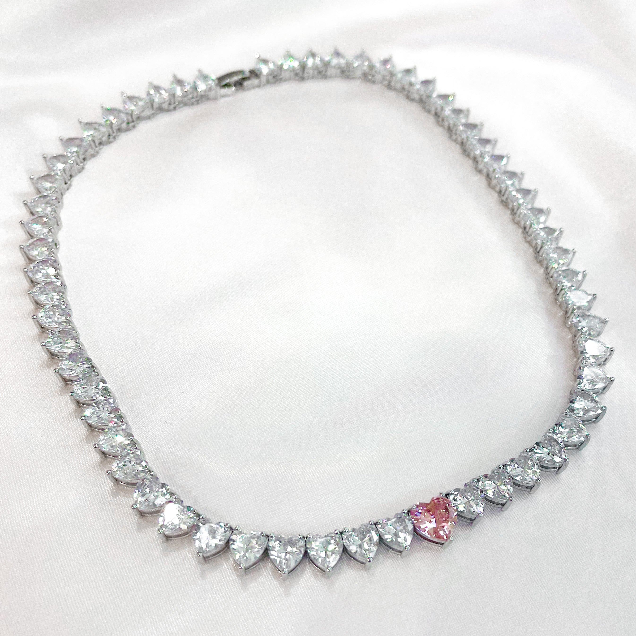 A sparkling silver-plated tennis-style necklace elegantly displayed on a soft, light background. It features a continuous row of heart-shaped cubic zirconia stones, each approximately 7 mm wide, set in rhodium-plated brass. The centerpiece is a single blush-pink heart stone (about 8 mm) that adds a pop of colour. The hearts are closely linked, creating a seamless chain around the neckline, and the full length is approximately 44 cm (17.5”) with a short extender. The polished silver setting enhances the bril