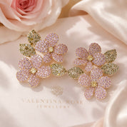 A pair of gold-tone floral stud earrings covered in pink and gold pavé stones, displayed on a cream satin surface with a pink rose and the Valentina Rose Jewellery logo.