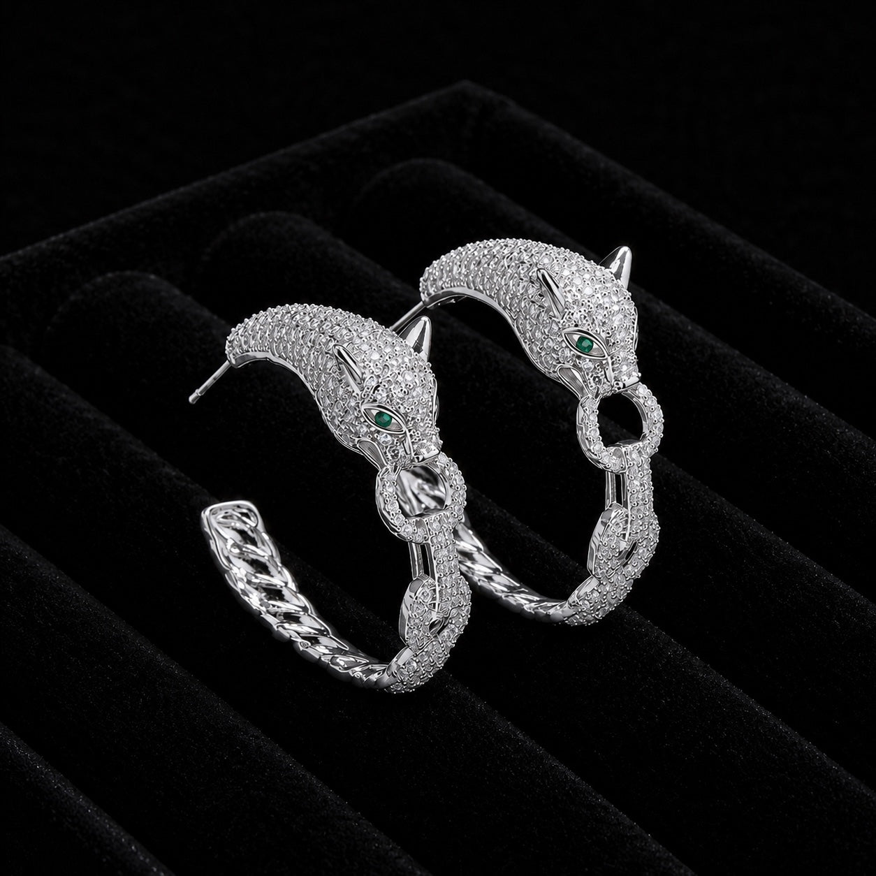 A pair of silver hoop earrings designed as stylized panther heads with small green eyes and paved with clear stones, displayed on a black velvet jewelry box.