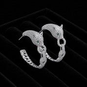 A pair of silver hoop earrings designed as stylized panther heads with small green eyes and paved with clear stones, displayed on a black velvet jewelry box.