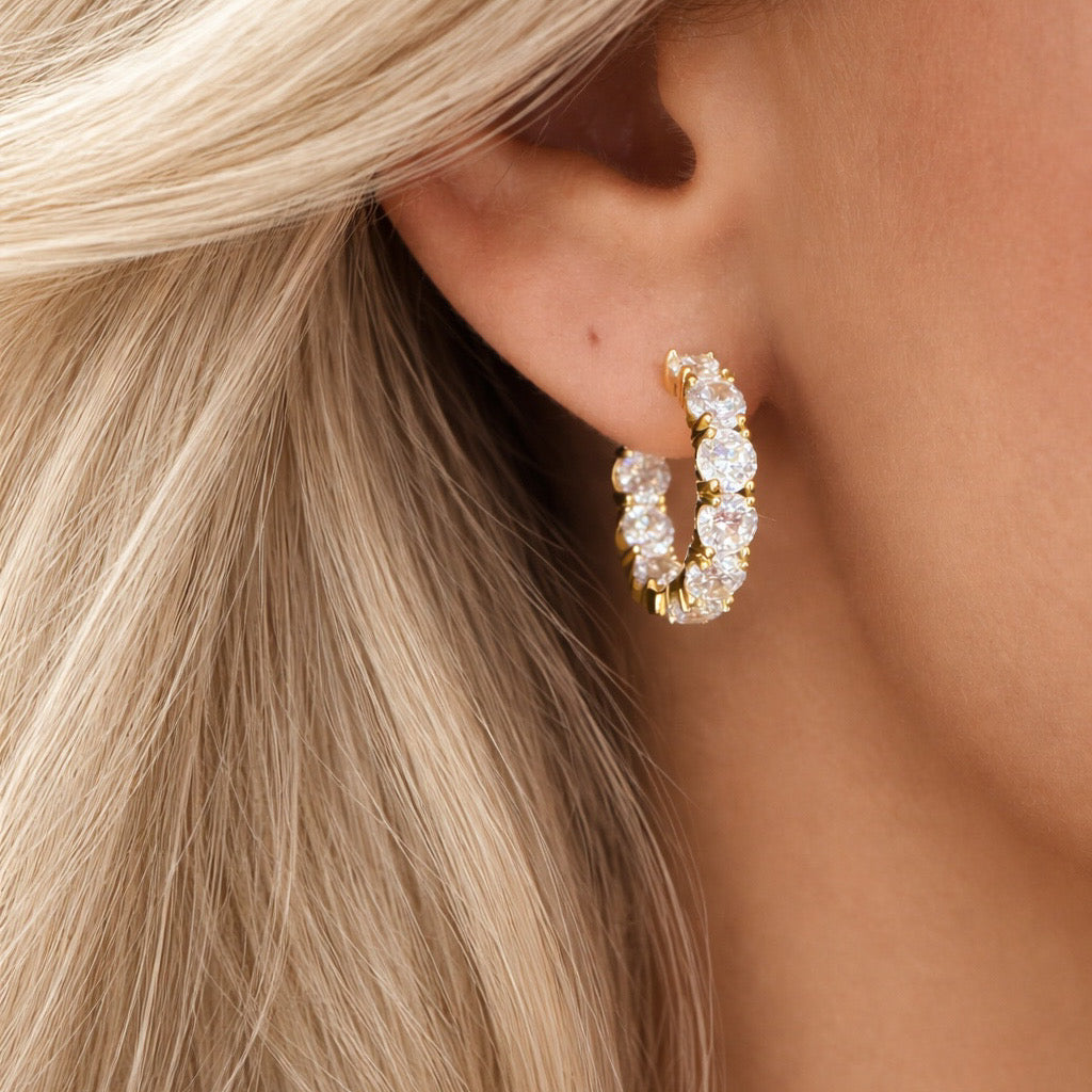 Close-up of a gold hoop earring with clear stones worn by a person with blonde hair.